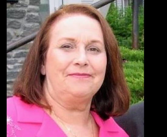 Lyn Simon, a woman with shoulder-length auburn hair, wearing a pink lapeled jacket, smiles at the camera.